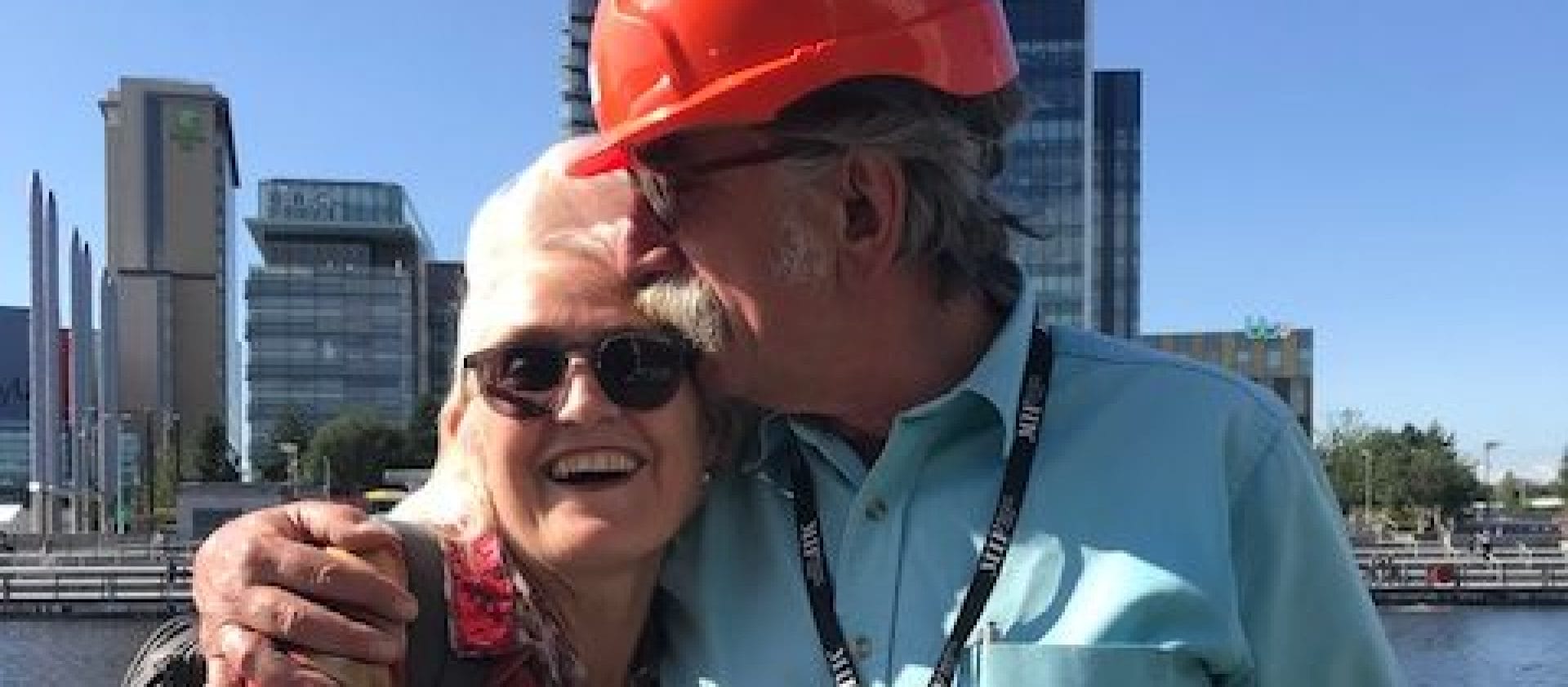 Rachel Clare in sunglasses and Simon Byford in a red hard hat stand together in a friendly embrace against a blue sky and tower blocks across the river We remember Simon Byford