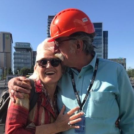 Rachel Clare in sunglasses and Simon Byford in a red hard hat stand together in a friendly embrace against a blue sky and tower blocks across the river We remember Simon Byford