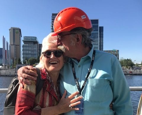 Rachel Clare in sunglasses and Simon Byford in a red hard hat stand together in a friendly embrace against a blue sky and tower blocks across the river We remember Simon Byford