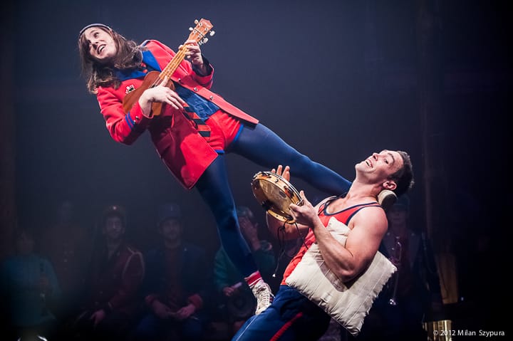 two performers in red and blue costumes in a dark circus tent with som musicians behind, one performer with a tambourine supports the other who plays a banjo