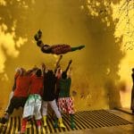 a group of performers in brightly pattered and colourful, mismatched clothes throw a performer into the air, ready to catch them, in front of a yellow sunlit wall.