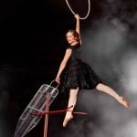 A performer in a black dress hanging gracefully from an aerial hoop above an iron board, set against a misty background.