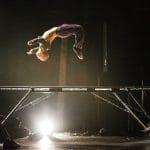 a performer in purple trousers and a grey shirt bounces on a trampoline turning themselves upside down, they are spotlit from below and two other performers sit on the floor either side watching