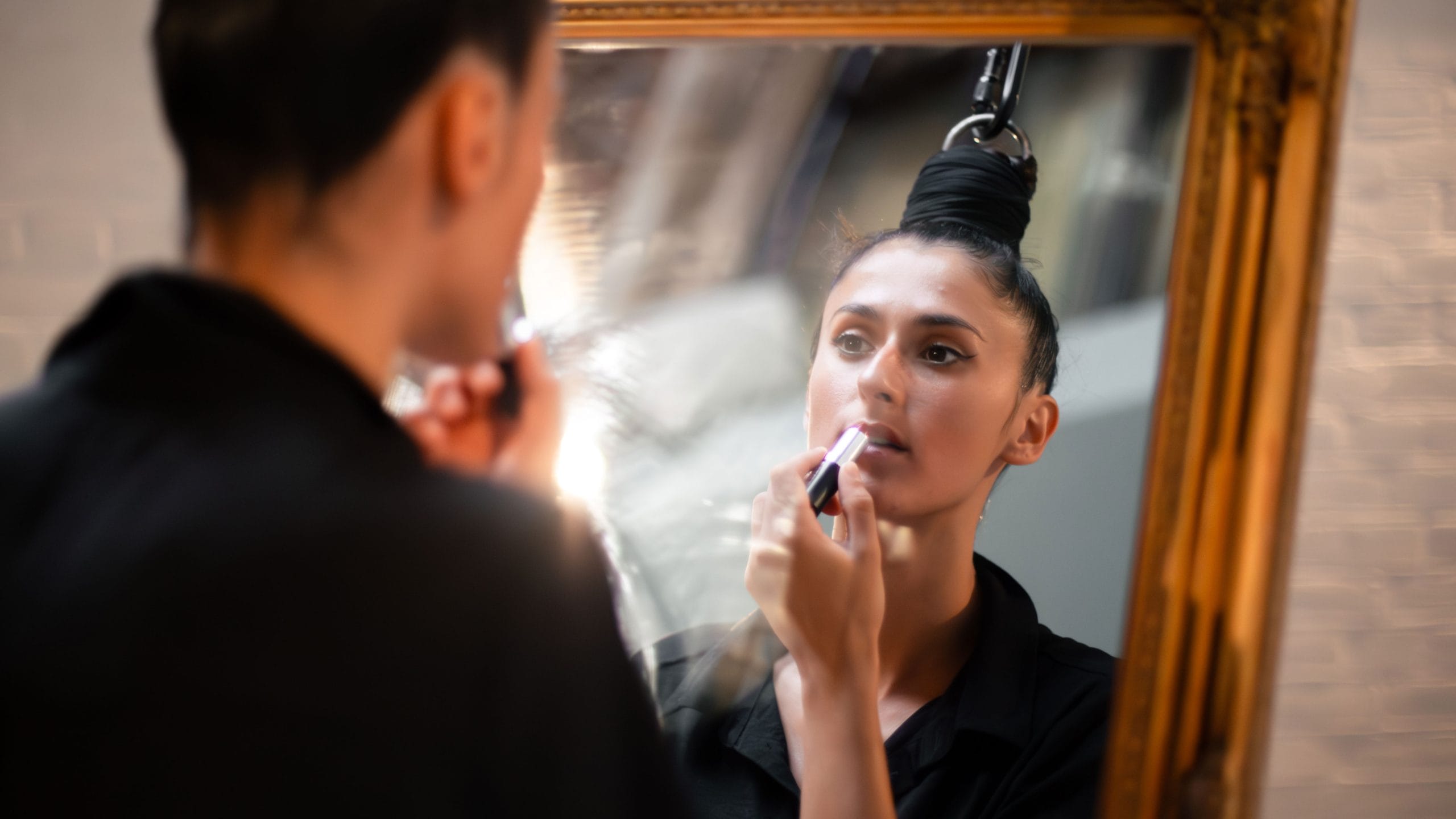 A person in a black shirt applies makeup in front of a vintage mirror, illuminated by soft light in a cosy setting.