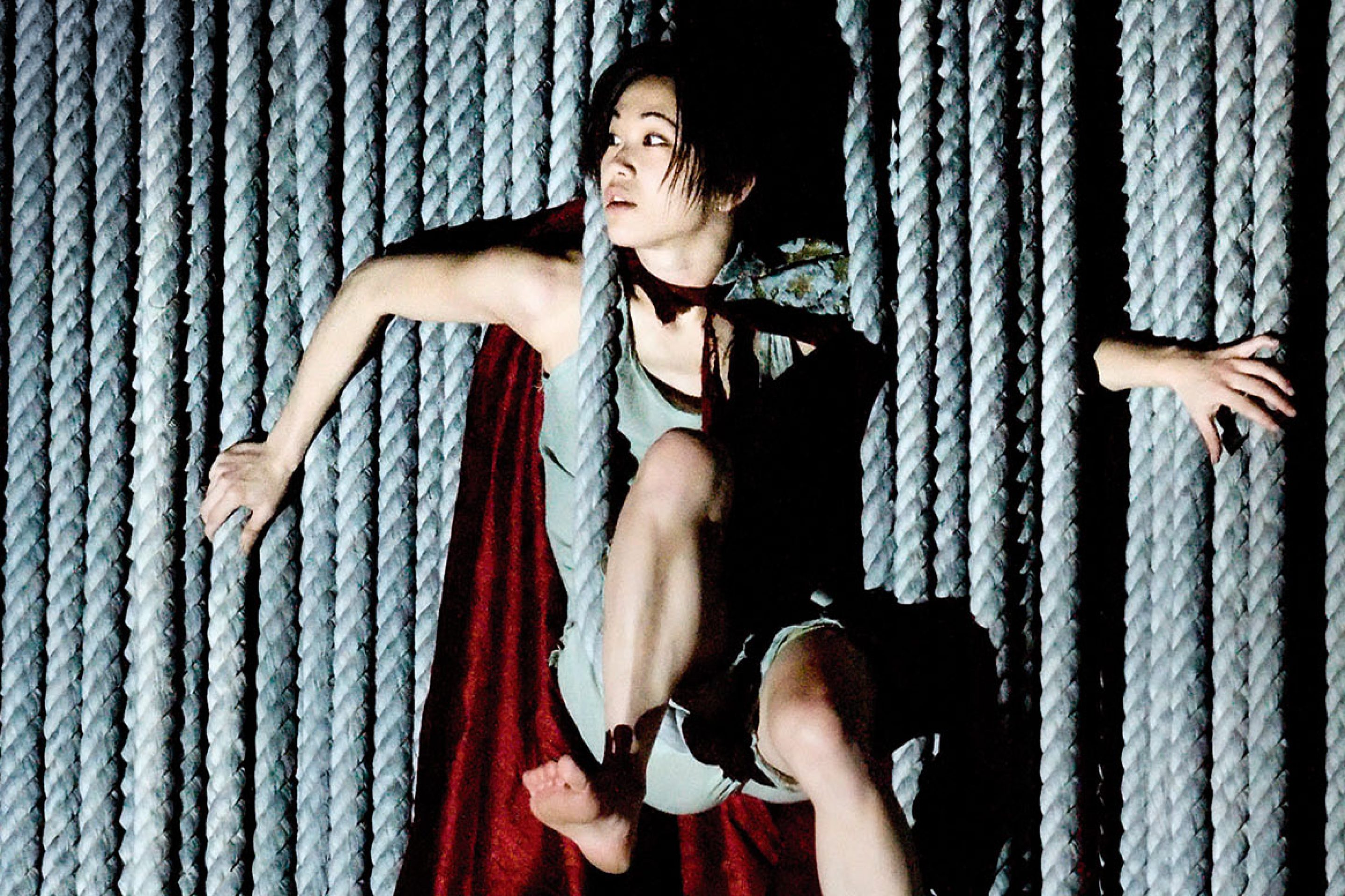 A tall, wide rope curtain hangs from top to bottom like a veil, revealing a performer emerging from between the rope strands, creating a unusual fragmented shapes. pushing boundaries contemporary performance