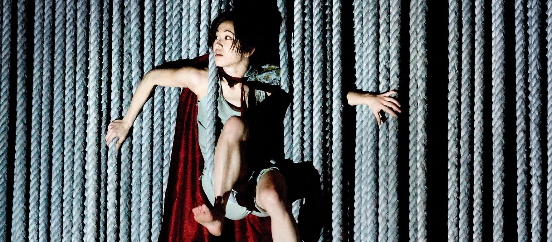 A tall, wide rope curtain hangs from top to bottom like a veil, revealing a performer emerging from between the rope strands, creating a unusual fragmented shapes. pushing boundaries contemporary performance
