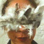 A close-up portrait of a person with a deep red colour lips, wearing an elaborate mask made of white and dark feathers, which completely covers their eyes, creating a striking, artistic look against a textured white background. Jeanne Mordoj Residencies