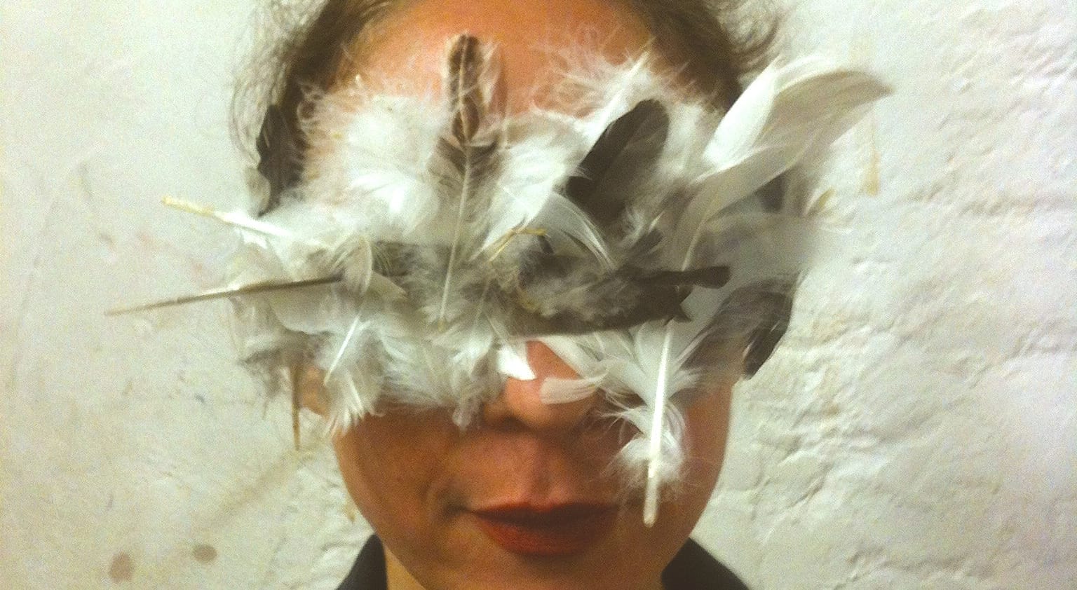 A close-up portrait of a person with a deep red colour lips, wearing an elaborate mask made of white and dark feathers, which completely covers their eyes, creating a striking, artistic look against a textured white background. Jeanne Mordoj Residencies