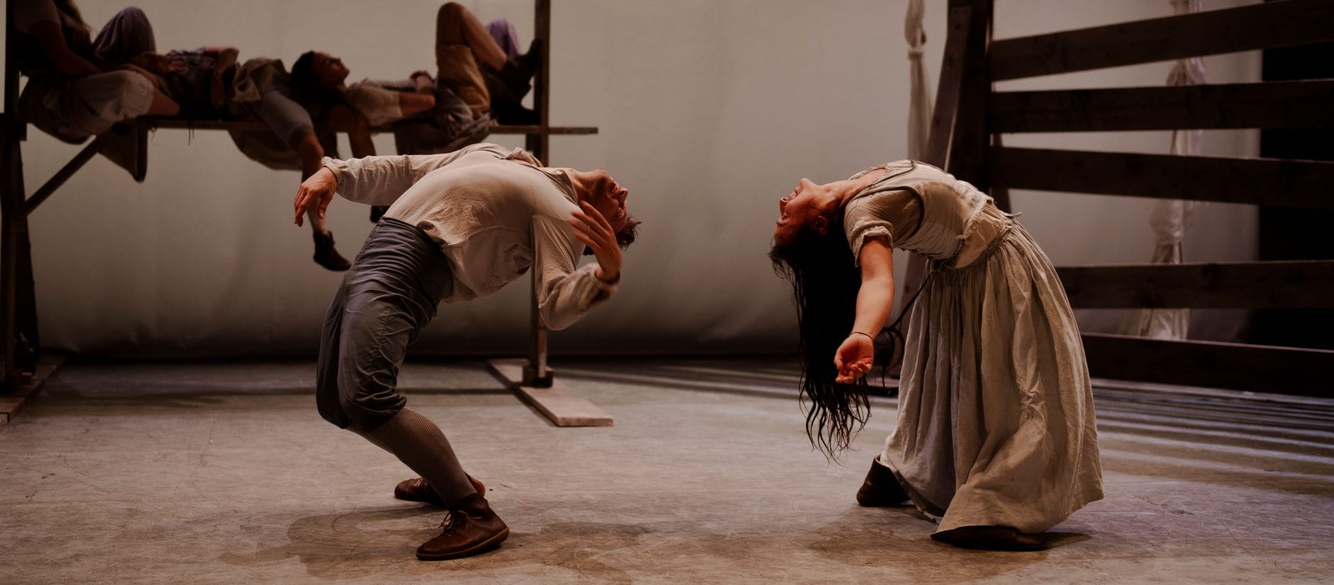 two performers on a stage stand side on to the audience, they wear dull pale brown clothes, one in trousers and a shirt, the other has long dark hair and a long dress, each bend over, nearly backwards, with their heads close to each other and their arms out at their sides