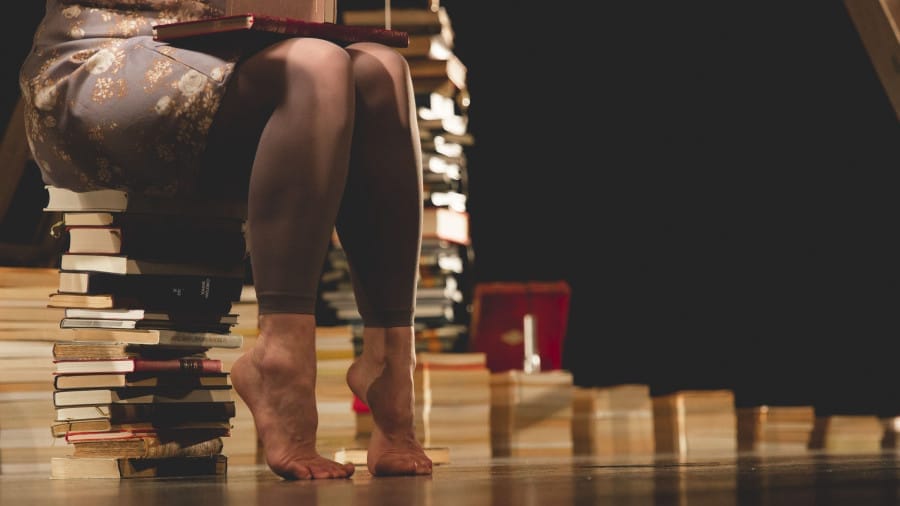 A woman with bare feet sits on a stack of books, holding a closed book in her lap, surrounded by more books in a dimly lit room.