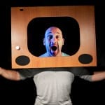 a bald headed, bearded performer with their hands out either side of them wears an old fashioned wooden television set frame on their head as if they are on television Atelier Lefeuvre André