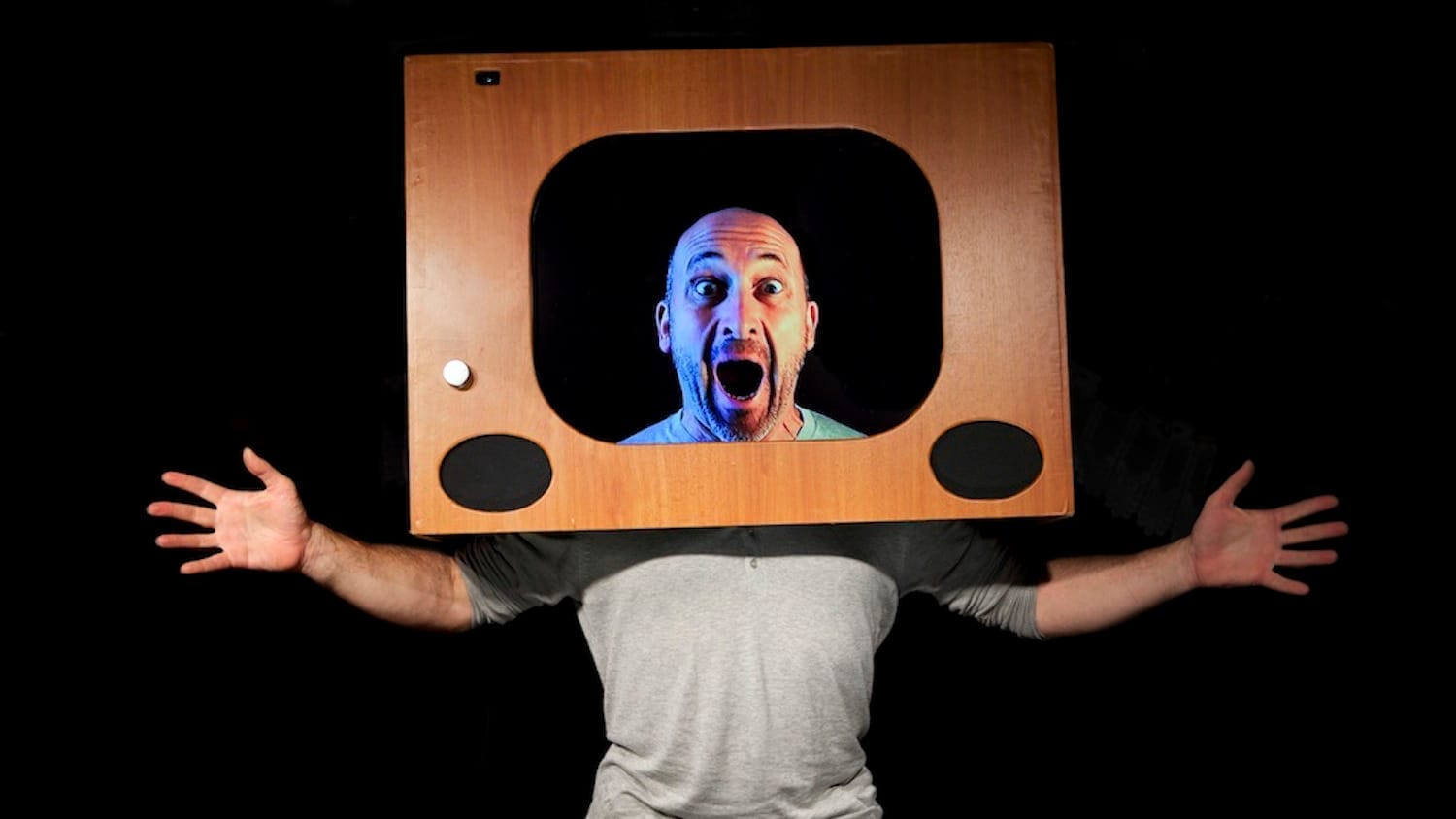 a bald headed, bearded performer with their hands out either side of them wears an old fashioned wooden television set frame on their head as if they are on television Atelier Lefeuvre André