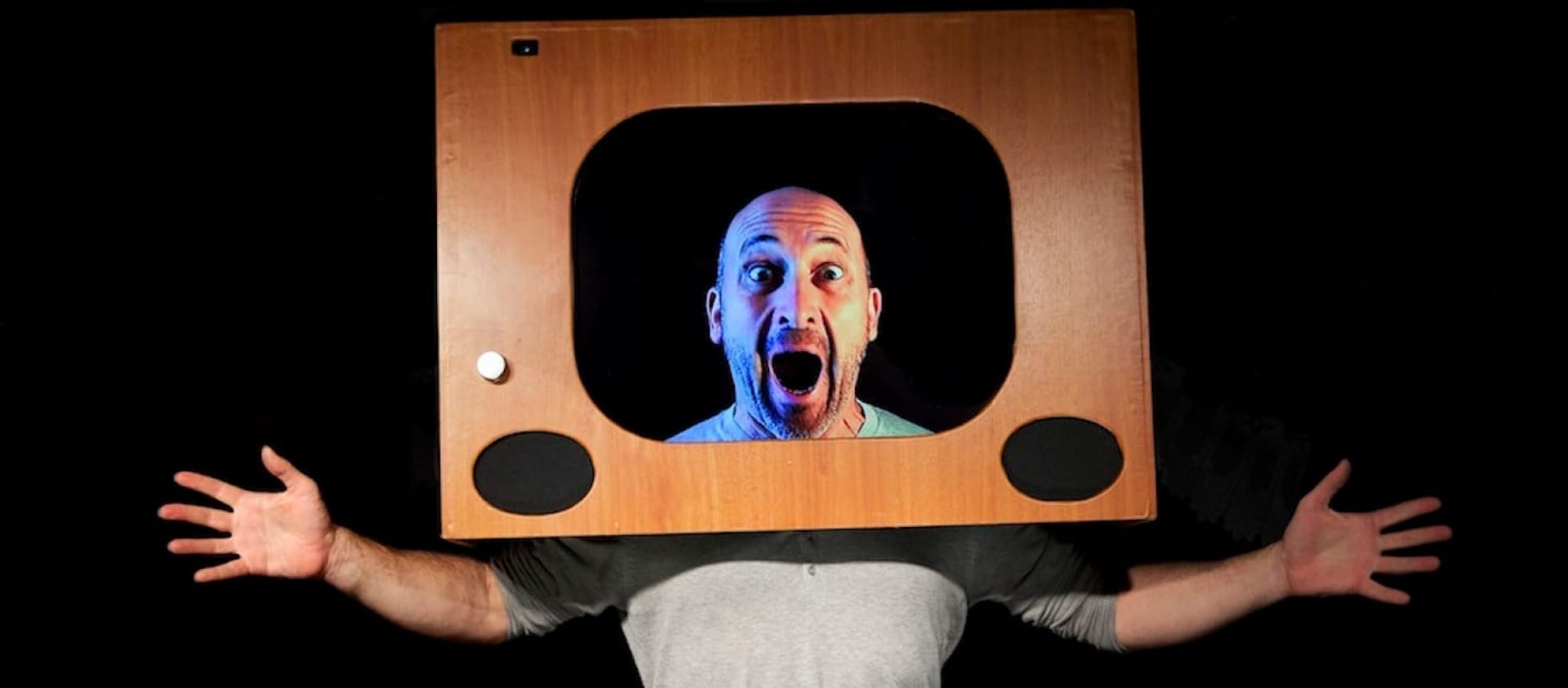 a bald headed performer with their hands out either side of them wears an old fashioned wooden television set frame on their head as if they are on television Atelier Lefeuvre André