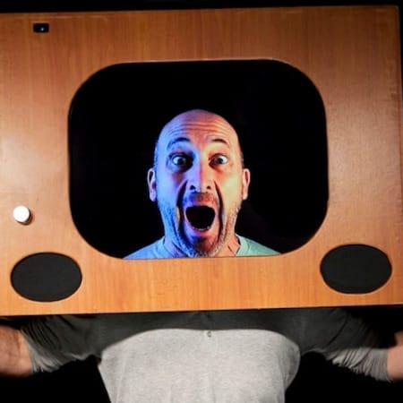 a bald headed performer with their hands out either side of them wears an old fashioned wooden television set frame on their head as if they are on television Atelier Lefeuvre André