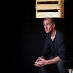 a shaven headed, performer with their hands clasped in front of them wears a blue buttoned t-shirt and dark jeans and sits on a wooden stool balancing a wooden crate on their head in a dark stage set Atelier Lefeuvre André