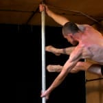 an acrobat wearing black underwear swings round athletically on a pole in a large but confined wooden crate, they have closely shaved hair and skin red from exertion Atelier LeFeuvre et André
