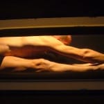a performer wearing black underwear is bent double in a confined metal space, they have closely shaved hair and are lit brightly Atelier LeFeuvre et André