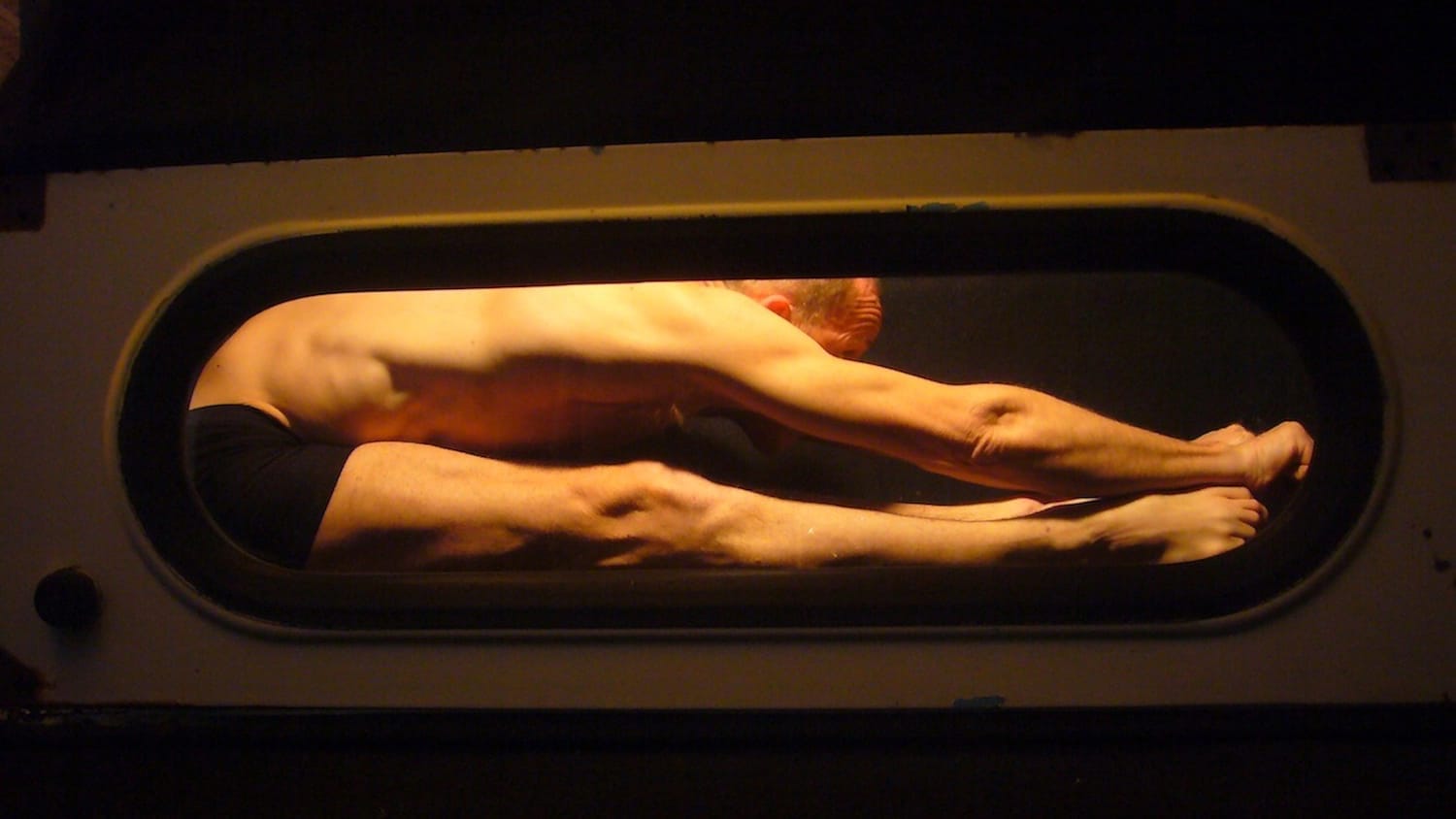 a performer wearing black underwear is bent double in a confined metal space, they have closely shaved hair and are lit brightly Atelier LeFeuvre et André