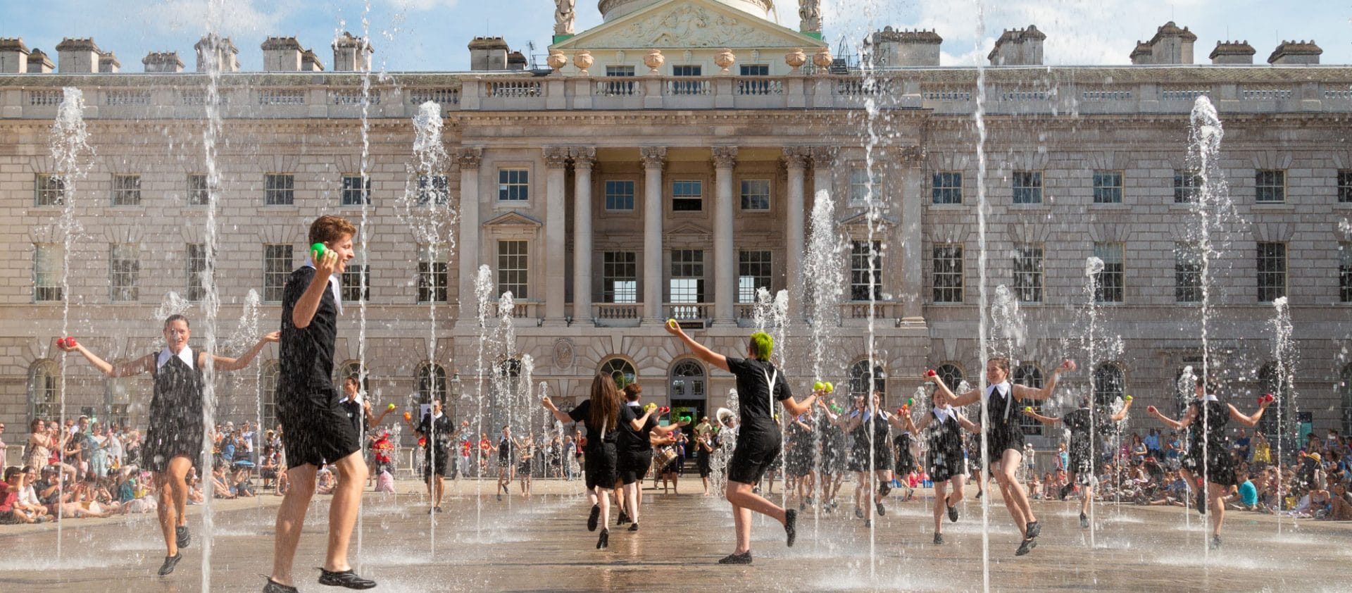 Performers juggling and dancing among fountains in front of a historic building, with a crowd watching in the background.
