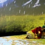 Three performers in colourful outfits interact artistically on a stage floor against a vibrant, projected landscape in the background with falling rain.