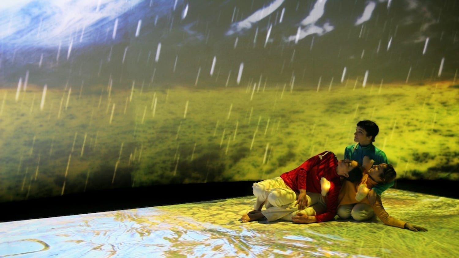 Three performers in colourful outfits interact artistically on a stage floor against a vibrant, projected landscape in the background with falling rain.