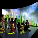 Group of children gathered in an immersive space, surrounded by vibrant projections of grass and clouds, engaging with the interactive environment.