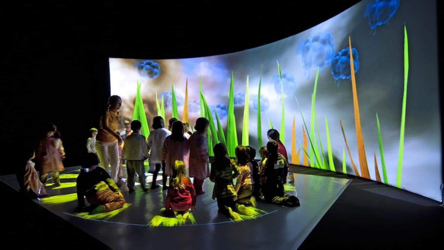 Group of children gathered in an immersive space, surrounded by vibrant projections of grass and clouds, engaging with the interactive environment.