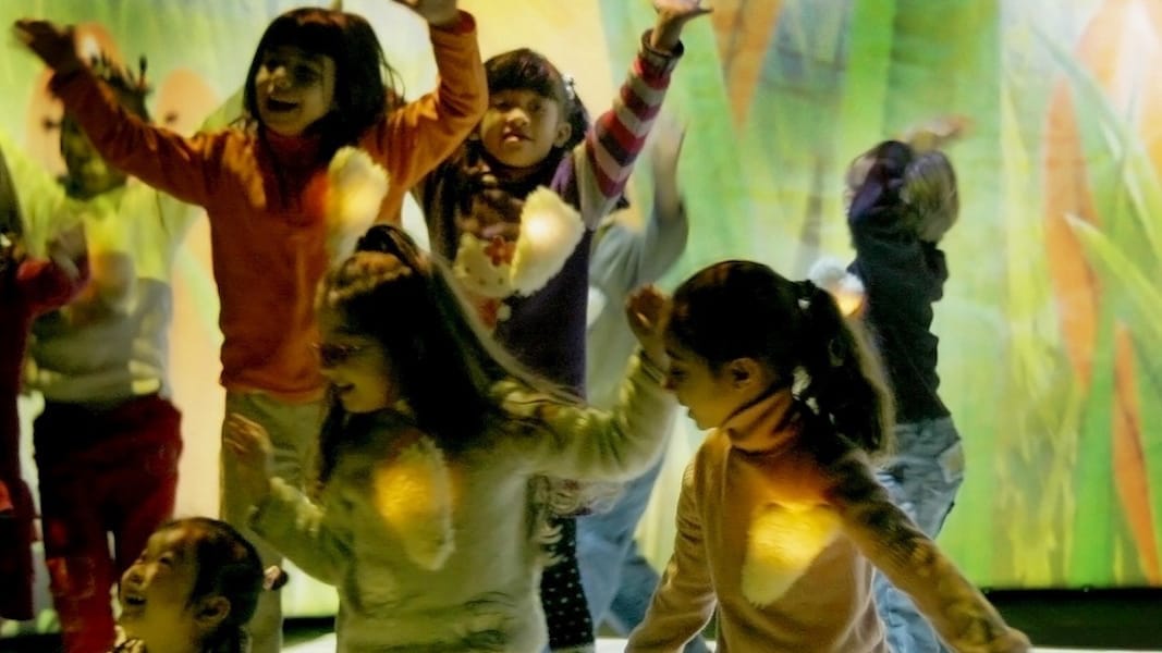 A lively group of children dancing joyfully against a colourful, nature-themed projection in an immersive space.