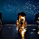 Three performers kneel on a dark reflective surface, surrounded by sparkling lights against a deep blue backdrop.