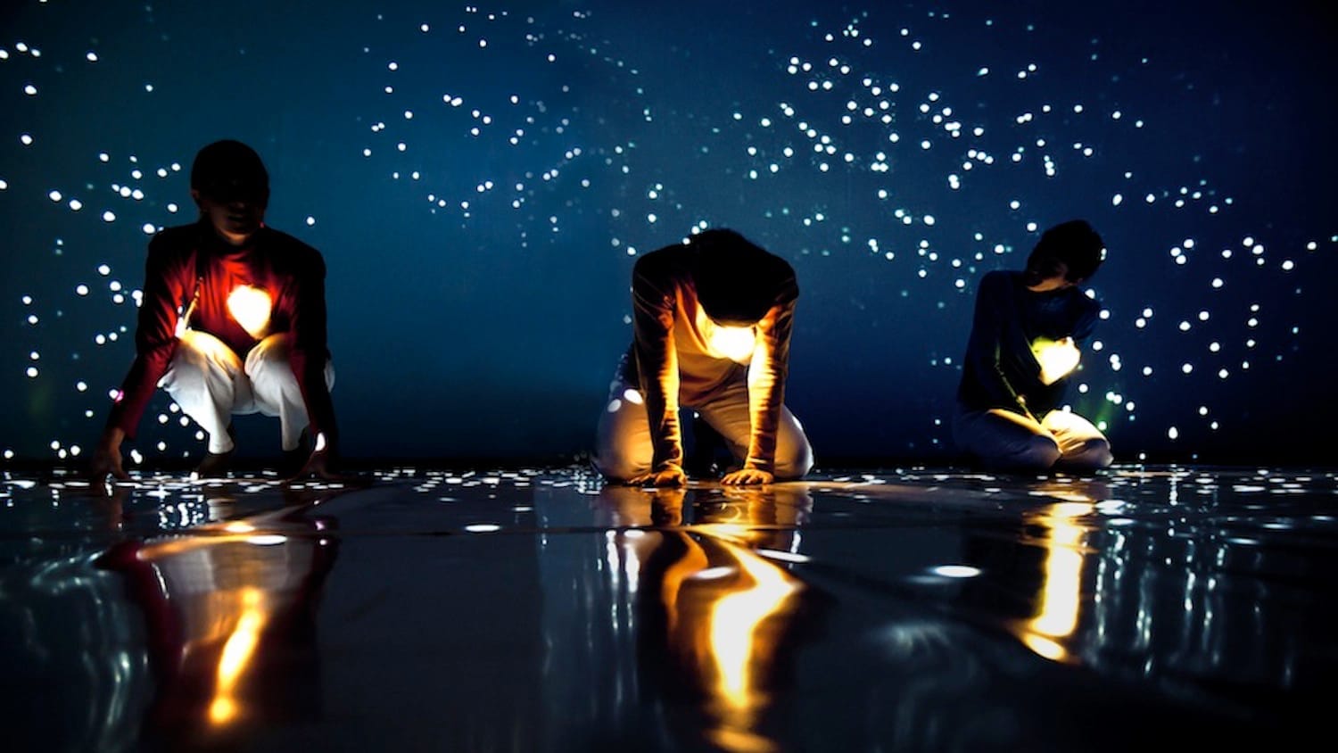 Three performers kneel on a dark reflective surface, surrounded by sparkling lights against a deep blue backdrop.
