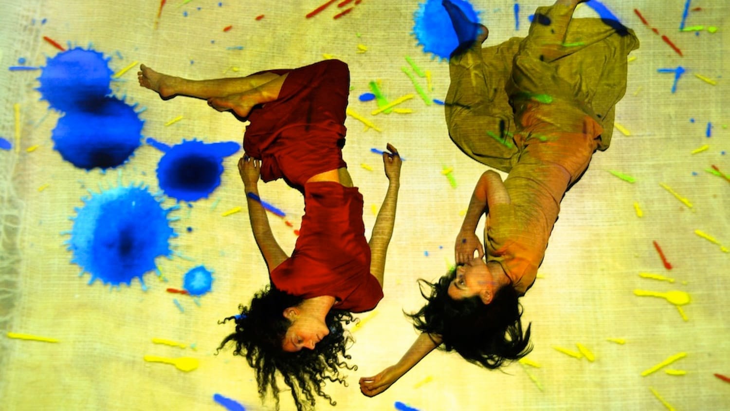 Two women in flowing red and yellow dresses lie on the ground, illuminated by a vibrant projection of canvas, surrounded by vibrant blue paint splatters.