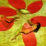 Two children lie on the ground, illuminated by a vibrant projection of a yellow canvas with a large red flower pattern, creating a dreamlike atmosphere.
