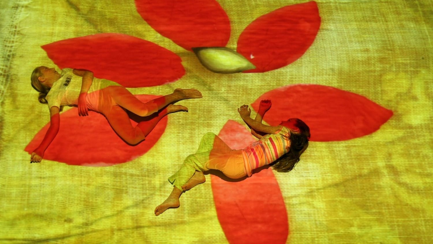 Two children lie on the ground, illuminated by a vibrant projection of a yellow canvas with a large red flower pattern, creating a dreamlike atmosphere.