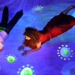 A person in a red dress and a child in a white shirt lie on a floor, illuminated by a vibrant blue projection with colourful patterns.