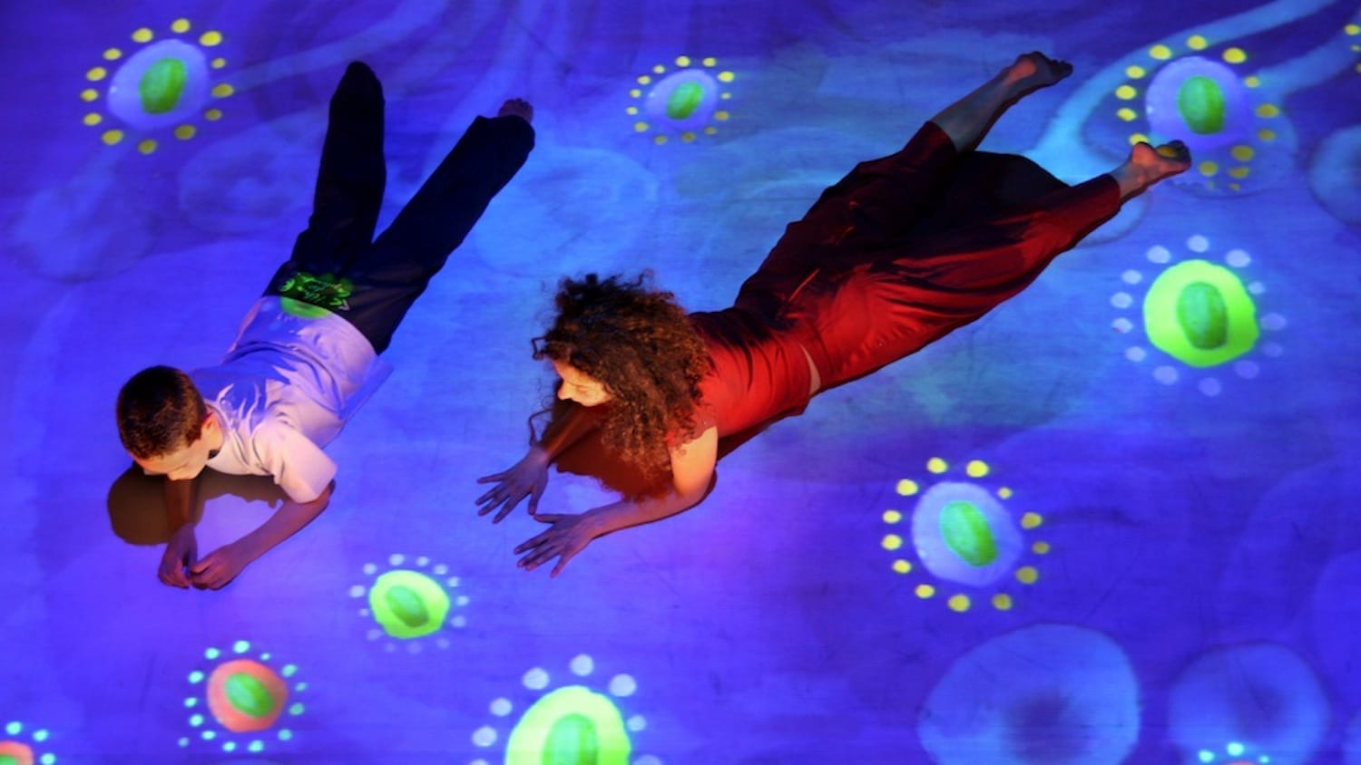 A person in a red dress and a child in a white shirt lie on a floor, illuminated by a vibrant blue projection with colourful patterns.