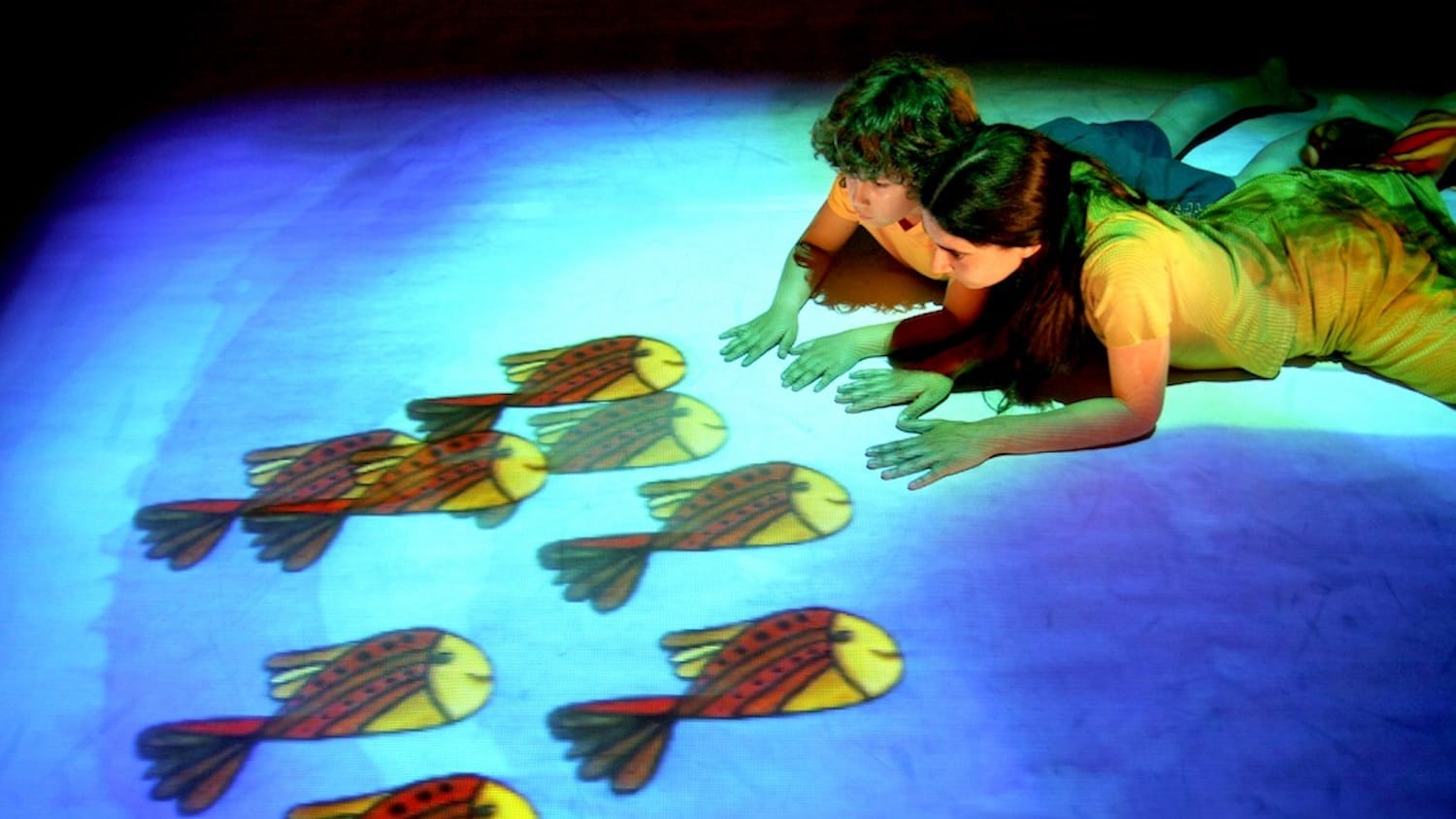 A child and an adult lie on the floor, fascinated by animated fish projected on the ground in vibrant colours.