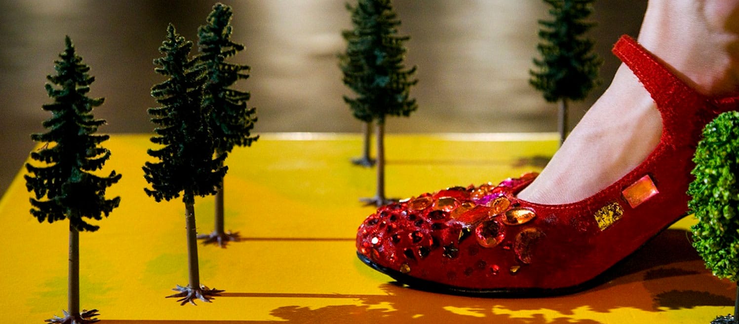 a close-up of a foot in a red shoe decorated with sparkling gems and sequinned embellishments, stepping on a yellow surface surrounded by miniature green fir trees resembling a small, crafted forest scene Dregs | inne Goris © Richard Haughton