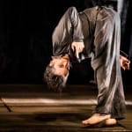 performer stands on stage barefoot in grey clothes, bending at the waist until they are bent double James Thierree Toad Knew