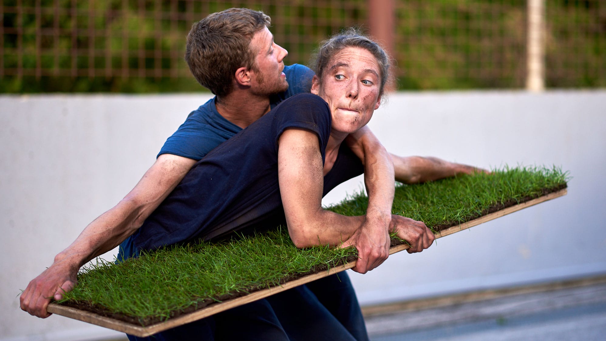 two performers in front of an outdoor street backdrop, one with long hair tied back and a navy blue t-shirt, the other with short brown hair and a lighter blue t-shirt, stand holding a table top with squares of grassy turf on it, they have muddy hands and arms and seems to fighting over the bright green turf