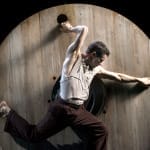 A person performs acrobatic moves on a large wooden wheel against a dark background, showcasing strength and agility. La Veillée des Abysses James Thierrée