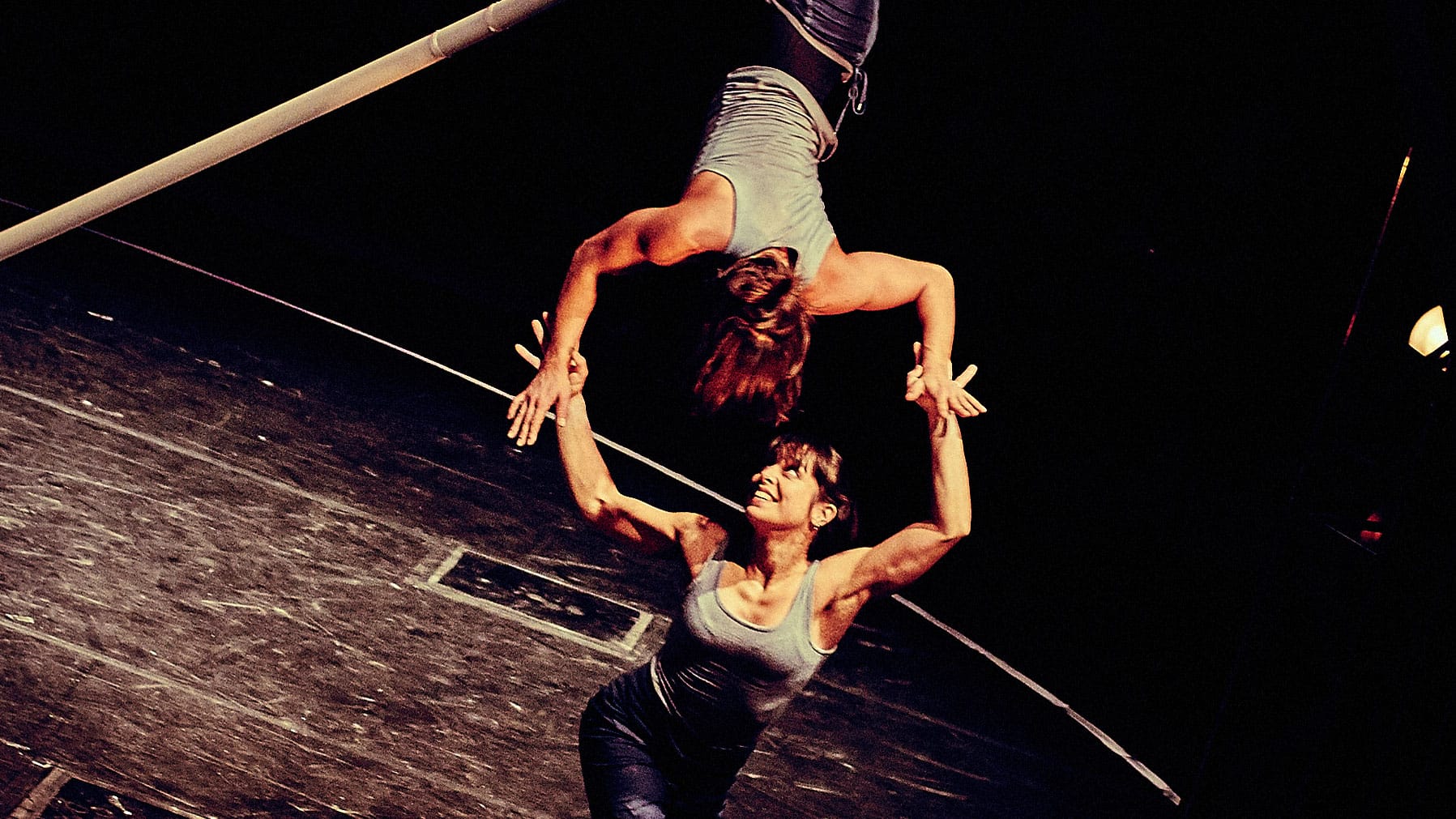 Two acrobats perform an aerial manoeuvre, one suspended upside down while the other extends their arms upwards. The moment freezes as they reach towards each other, their palms gently connecting in perfect synchronisation, showing both strength and elegance.