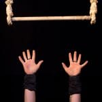 Two outstretched hands, wrapped in black wristbands, reach upwards towards a suspended wooden bar secured with rope, set against a dark background.