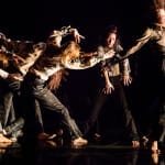 Seven performers on a dark indoor stage, all with long hair, dressed in a dark trousers and grey shirts move as one long queue of dance James Thierrée Tabac Rouge