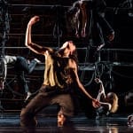 Performer kneels on an indoor stage, dressed in a dark brown trousers and a gold sleeveless top with long brown hair surrounded by other performers who hang their bodies, as if asleep, off bits of the stage set, all softly lit by blueish light James Thierrée Tabac Rouge