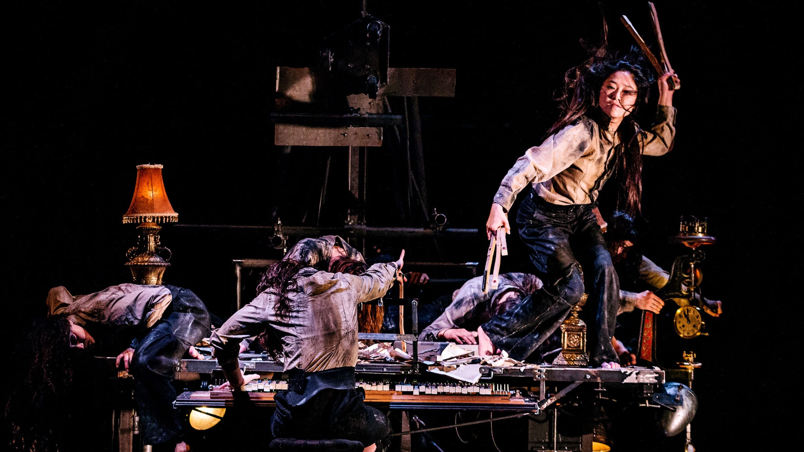 Performer stands on a messy desk on an indoor stage, dressed in a dark trousers and grey shirt and looks at the camera, surrounded by other performers dressed the same and softly lit by golden desk lamps James Thierrée Tabac Rouge