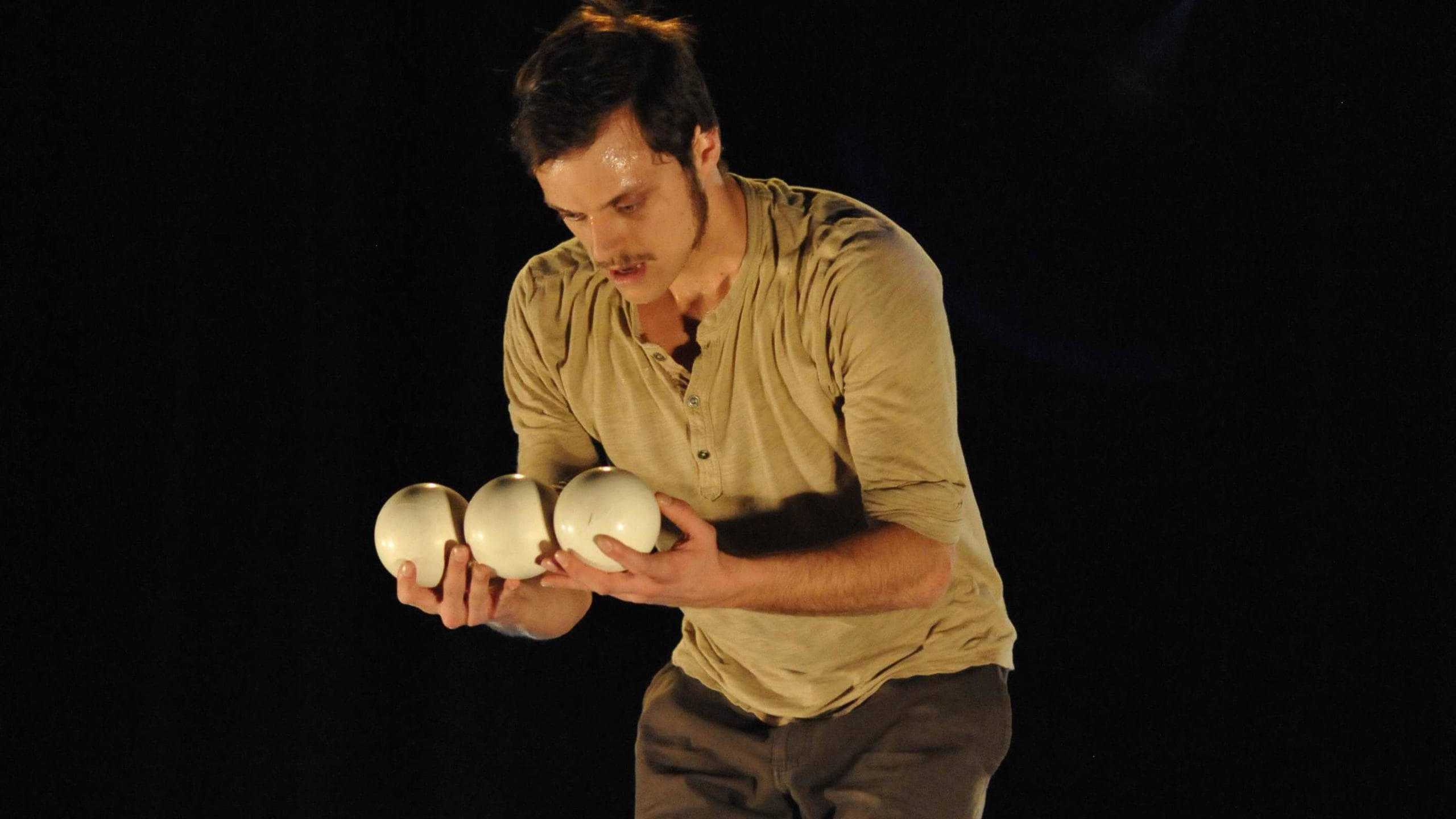 an acrobat with brown hair and beard, wearing brown trousers and grey sweatshirt performs with three juggling balls in an indoor stage setting with yellow lighting