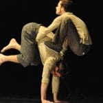 two acrobats both with brown hair and beards, wearing brown trousers and grey sweatshirts perform in an indoor stage setting with yellow lighting