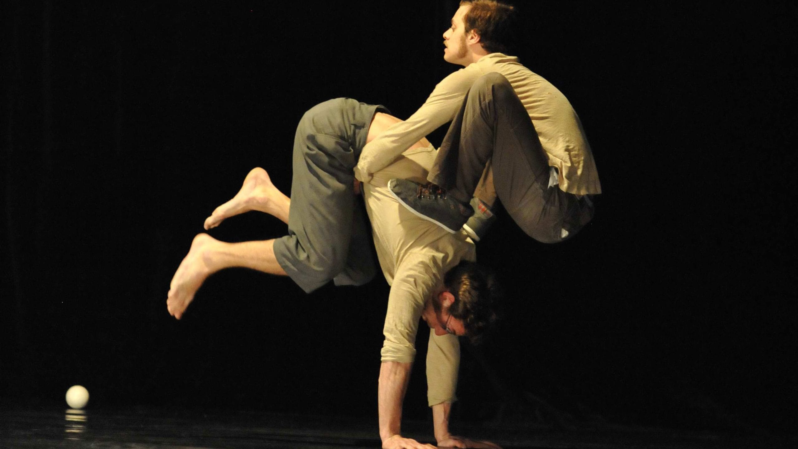 two acrobats both with brown hair and beards, wearing brown trousers and grey sweatshirts perform in an indoor stage setting with yellow lighting