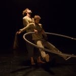 two acrobats both with brown hair and beards, wearing brown trousers and grey sweatshirts perform over a gym ball and with a cyr wheel in an indoor stage setting with yellow lighting