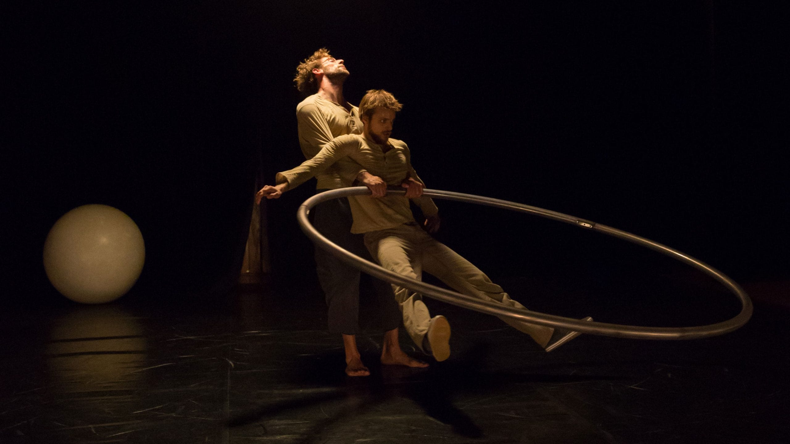 two acrobats both with brown hair and beards, wearing brown trousers and grey sweatshirts perform over a gym ball and with a cyr wheel in an indoor stage setting with yellow lighting
